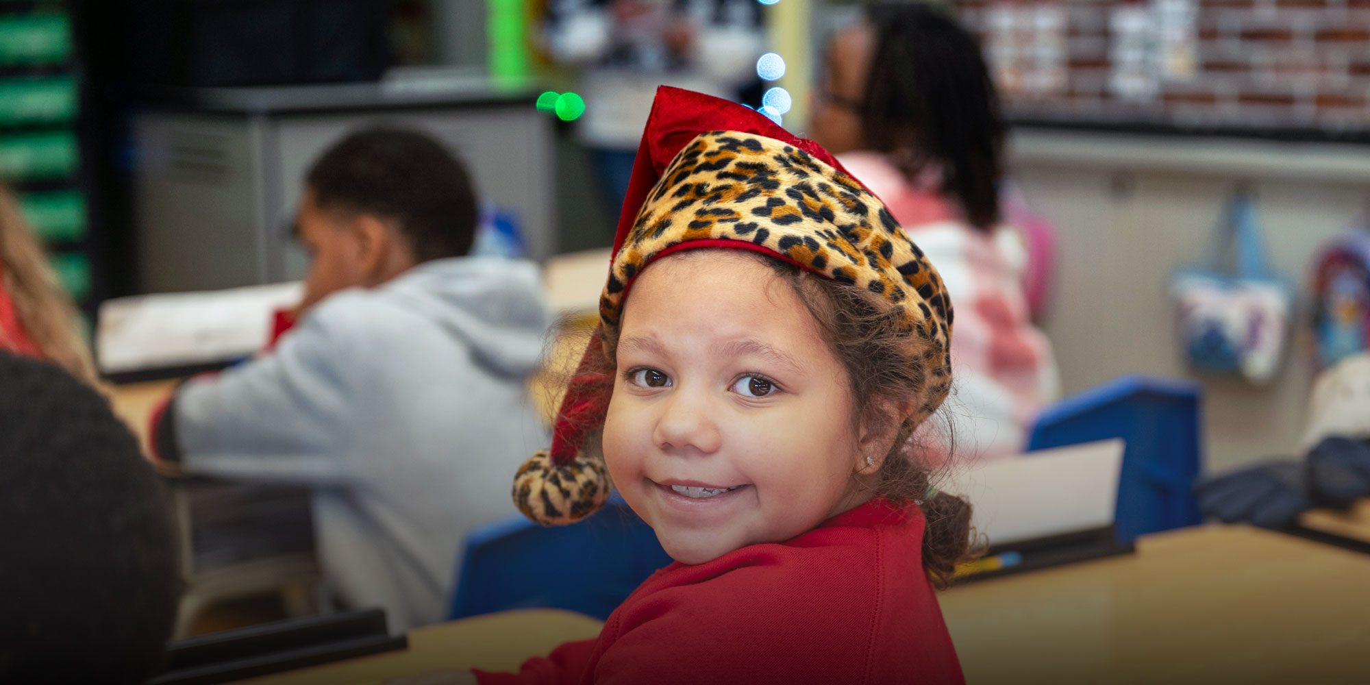 Smiling student in a leopard-print hat seated at a classroom desk.